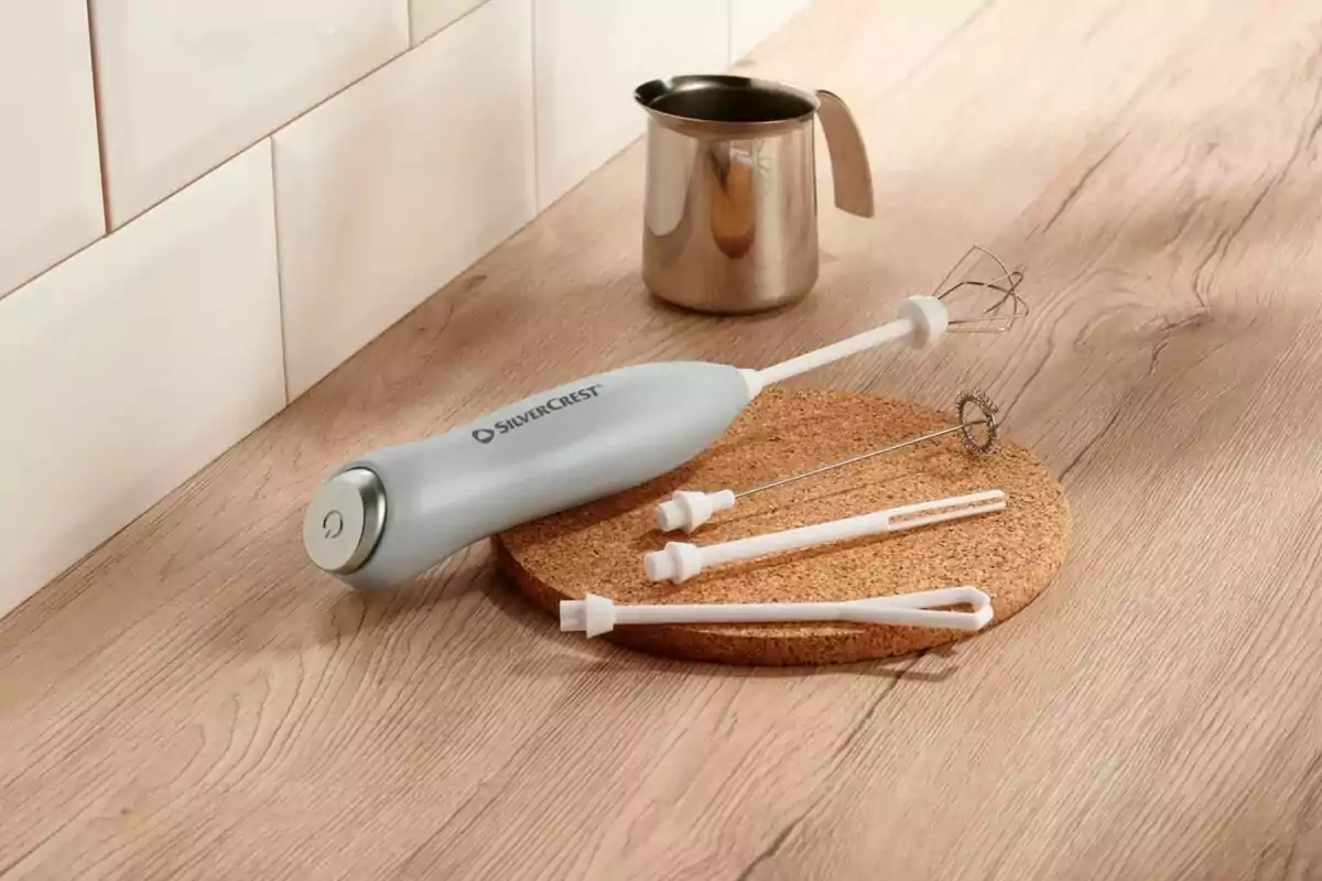 Batidora de mano eléctrica con varios accesorios sobre una base de corcho junto a una jarra metálica en una cocina Batidora de mano eléctrica con varios accesorios sobre una base de corcho junto a una jarra metálica en una cocina