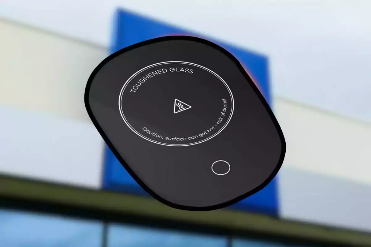 Dispositivo de inducción portátil de vidrio templado con advertencia de superficie caliente sostenido frente a un edificio desenfocado. Dispositivo de inducción portátil de vidrio templado con advertencia de superficie caliente sostenido frente a un edificio desenfocado.
