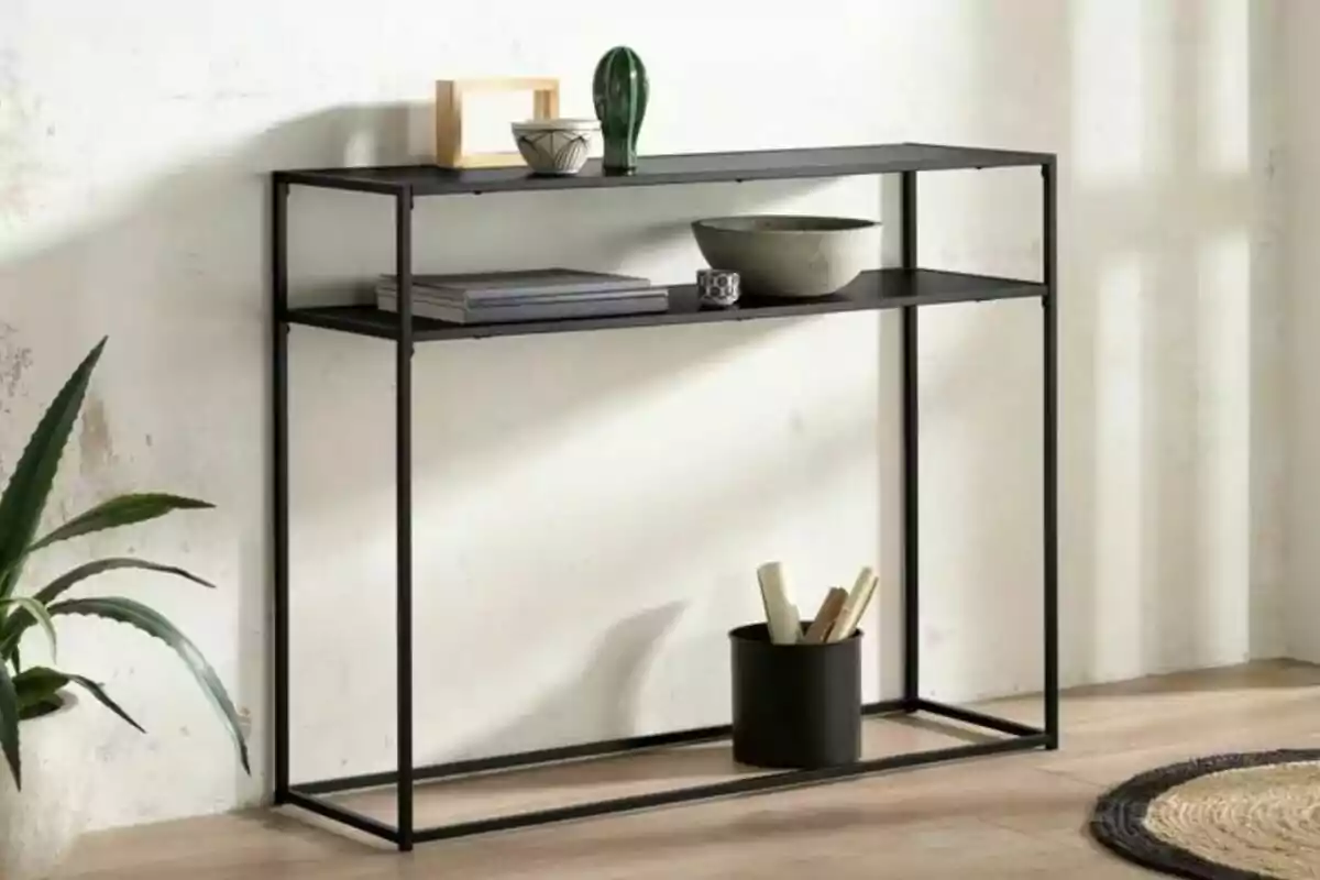 Mueble consola de metal negro con dos repisas decorado con libros, cuencos, una figura verde y una maceta al lado Mueble consola de metal negro con dos repisas decorado con libros, cuencos, una figura verde y una maceta al lado