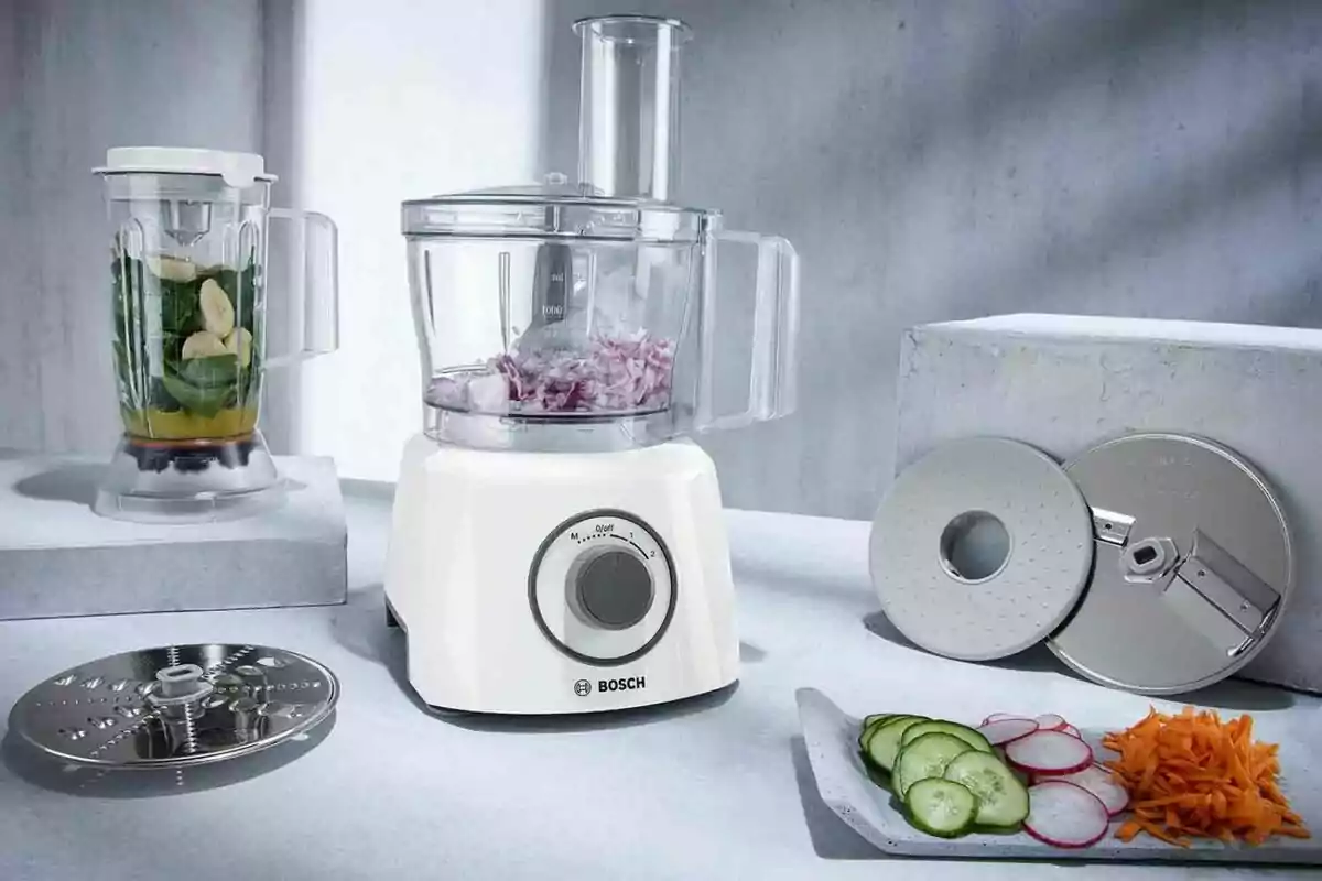 Procesador de alimentos blanco con accesorios metálicos, un vaso de licuadora con frutas y verduras, y un plato con rodajas de pepino, rábano y zanahoria rallada Procesador de alimentos blanco con accesorios metálicos, un vaso de licuadora con frutas y verduras, y un plato con rodajas de pepino, rábano y zanahoria rallada