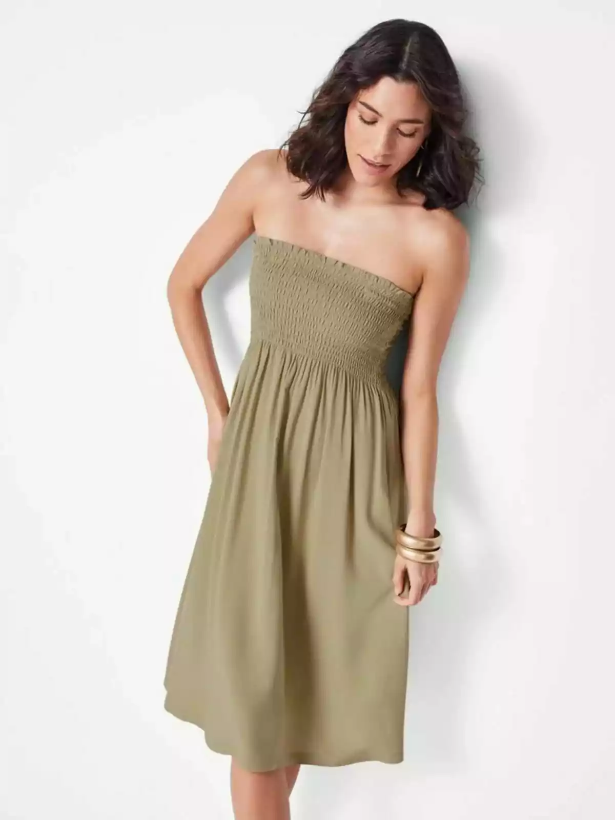Mujer con cabello oscuro y ondulado usando un vestido strapless color verde oliva y pulseras grandes, posando de pie junto a una pared blanca Mujer con cabello oscuro y ondulado usando un vestido strapless color verde oliva y pulseras grandes, posando de pie junto a una pared blanca