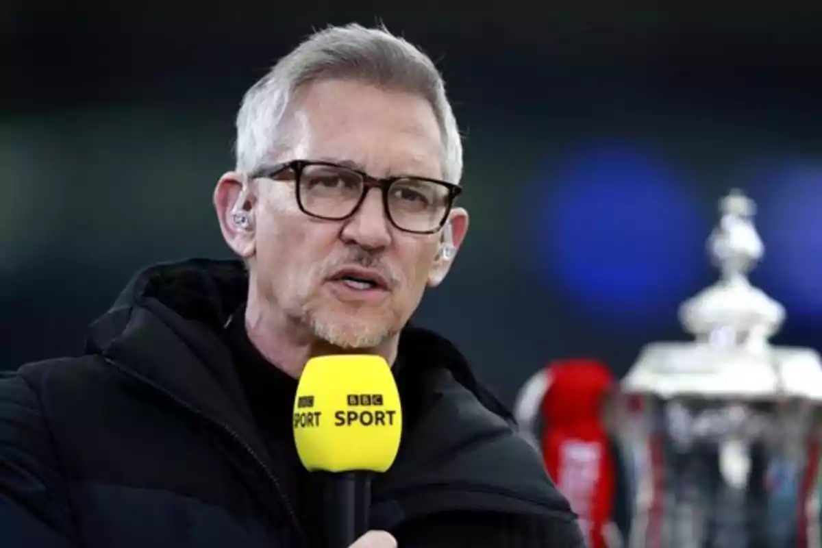 Hombre de cabello canoso y gafas hablando frente a un micrófono amarillo de BBC Sport con un trofeo plateado al fondo