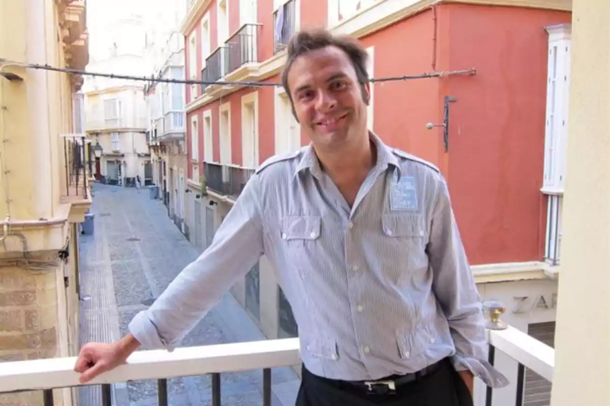 Hombre sonriente apoyado en la barandilla de un balcón en una calle estrecha con edificios de colores claros y rojizos Hombre sonriente apoyado en la barandilla de un balcón en una calle estrecha con edificios de colores claros y rojizos