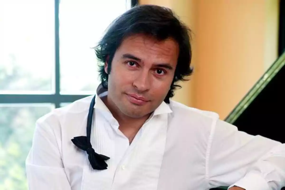 Hombre de cabello oscuro y camisa blanca con moño desatado sentado frente a una ventana