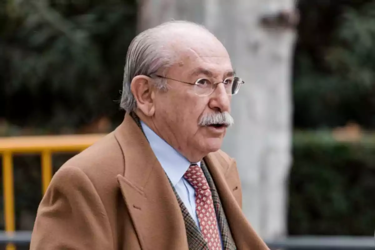 Hombre mayor con bigote y gafas usando abrigo marrón y corbata caminando al aire libre