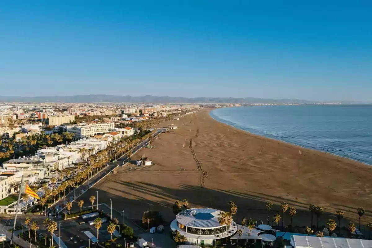 Vista aérea de una playa amplia con arena dorada junto a una ciudad costera y el mar en calma bajo un cielo despejado Vista aérea de una playa amplia con arena dorada junto a una ciudad costera y el mar en calma bajo un cielo despejado
