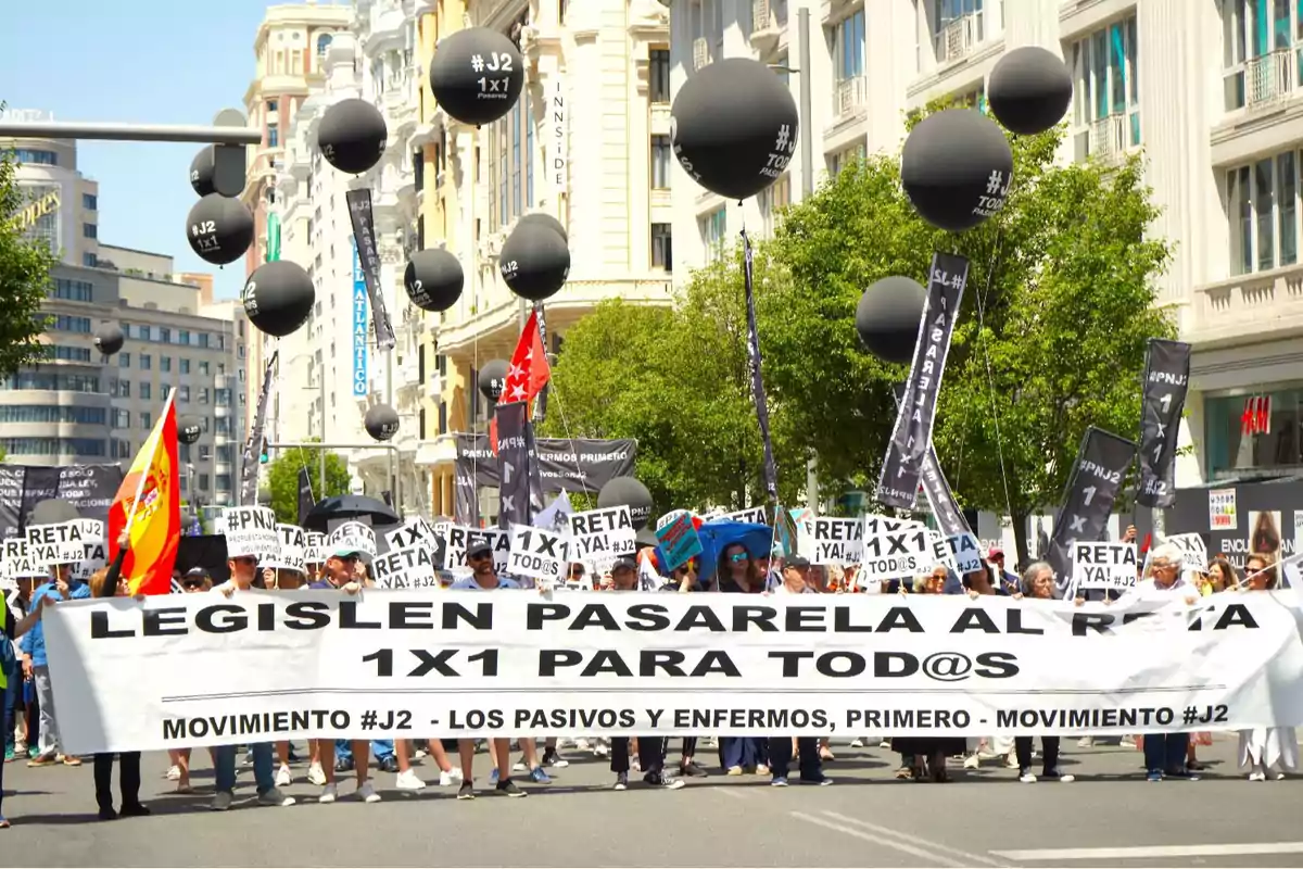 Manifestación en una avenida con personas sosteniendo una pancarta grande que pide legislar la pasarela al RETA 1x1 para todos, acompañados de globos negros y banderas, en un entorno urbano con edificios y árboles. Manifestación en una avenida con personas sosteniendo una pancarta grande que pide legislar la pasarela al RETA 1x1 para todos, acompañados de globos negros y banderas, en un entorno urbano con edificios y árboles.