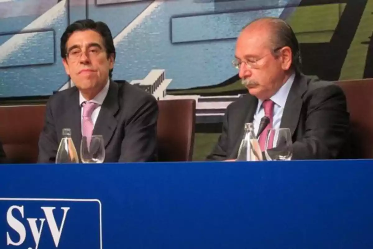 Dos hombres con traje y corbata sentados en una mesa azul durante una conferencia de prensa
