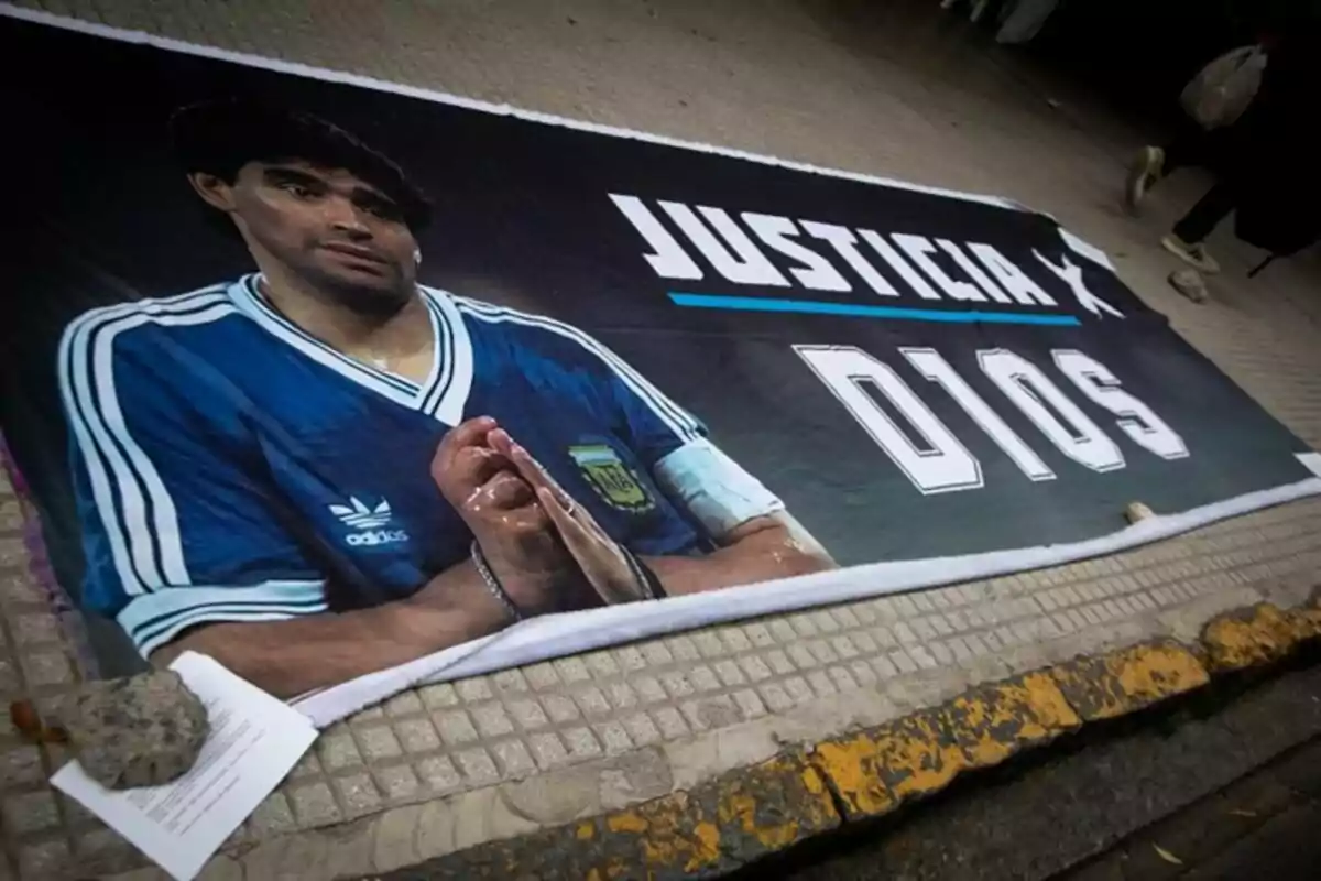 Pancarta en el suelo con la imagen de un futbolista argentino con las manos juntas y la palabra justicia junto a la palabra D10S
