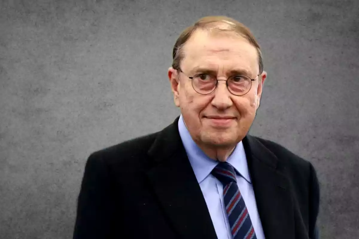 Hombre mayor con gafas y traje oscuro sobre fondo gris