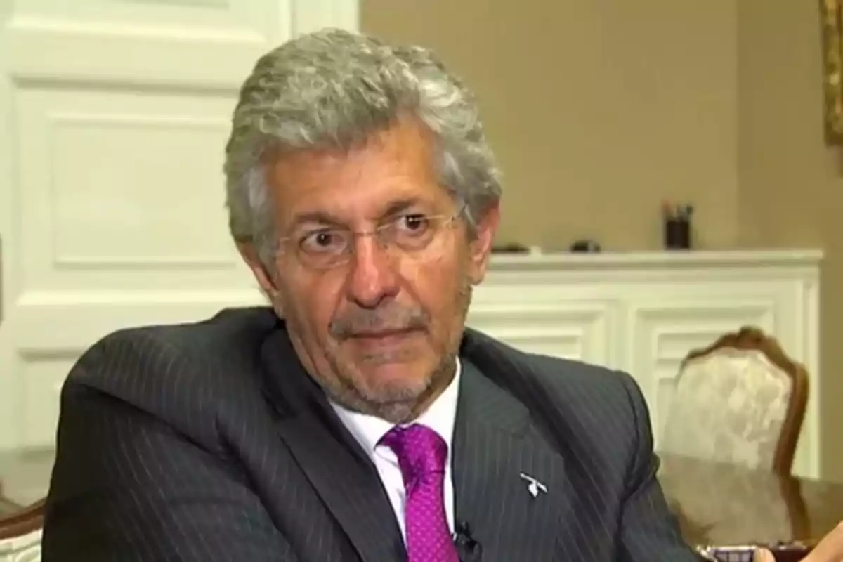 Hombre de cabello canoso y gafas con traje oscuro y corbata fucsia sentado en una oficina elegante
