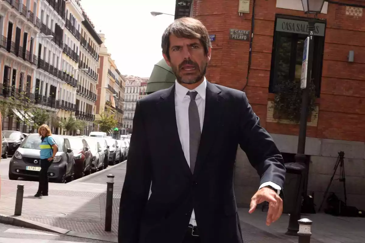 Hombre con traje oscuro y corbata caminando por una calle urbana con edificios y coches estacionados al fondo