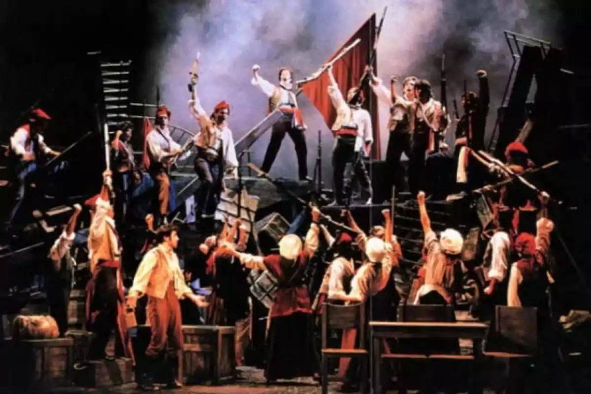 Un grupo de actores en el escenario representa una escena de revolución con banderas y armas improvisadas mientras levantan los brazos en señal de lucha Un grupo de actores en el escenario representa una escena de revolución con banderas y armas improvisadas mientras levantan los brazos en señal de lucha
