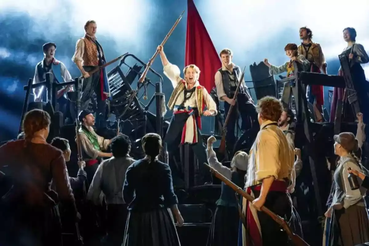 Un grupo de actores en el escenario representa una escena revolucionaria con vestuario de época y una bandera roja al fondo