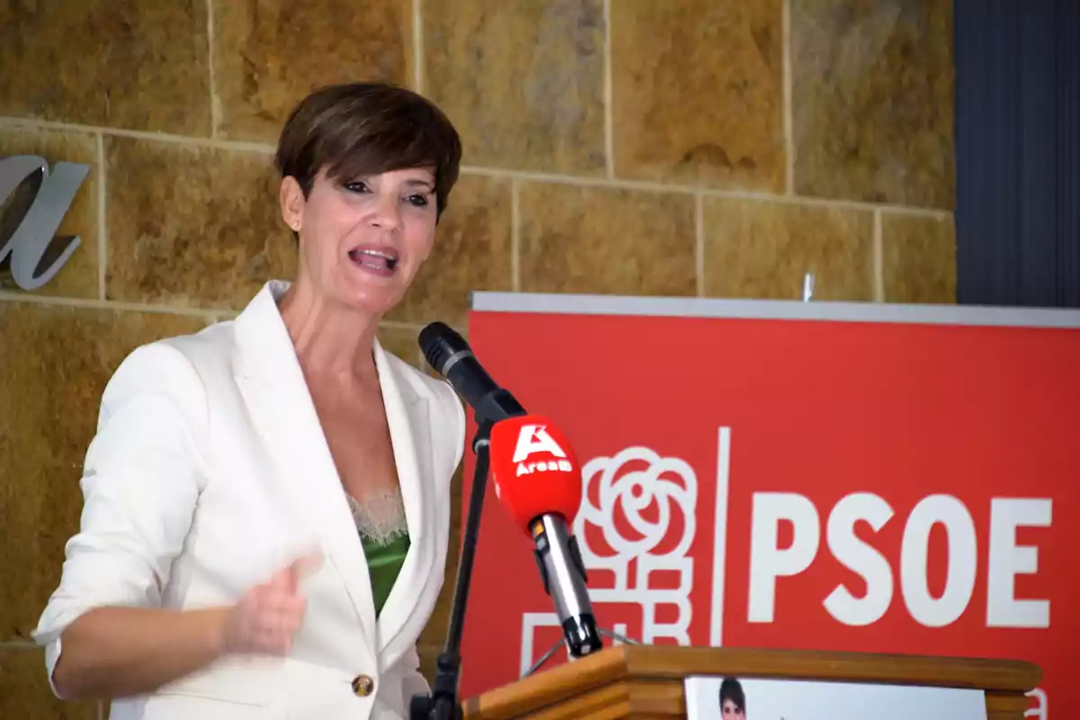 Mujer con chaqueta blanca hablando en un atril con micrófono frente a un cartel del PSOE
