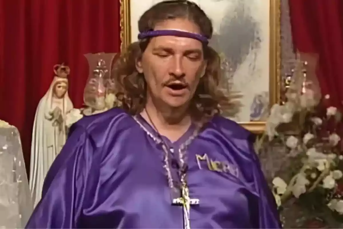 Hombre con túnica morada y cinta en la cabeza, rodeado de imágenes religiosas y flores, con un crucifijo colgado al cuello Hombre con túnica morada y cinta en la cabeza, rodeado de imágenes religiosas y flores, con un crucifijo colgado al cuello