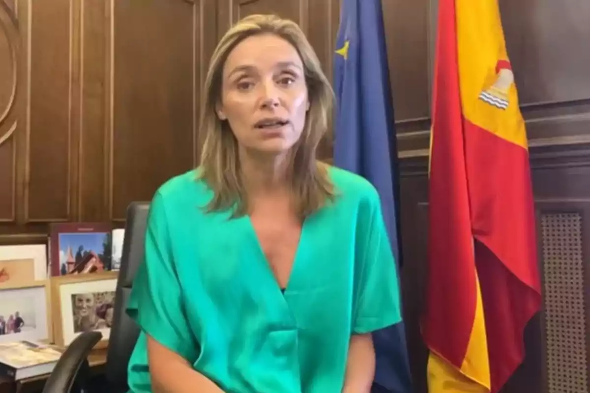 Mujer rubia con blusa verde sentada en una oficina con banderas de España y la Unión Europea al fondo