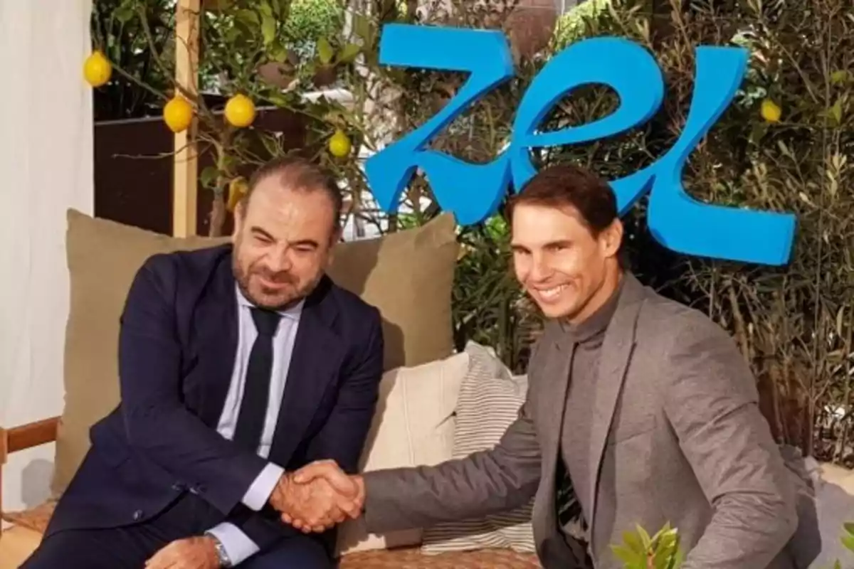 Dos hombres vestidos de traje se dan la mano y sonríen mientras están sentados en un ambiente decorado con plantas y limones, con un letrero azul grande al fondo Dos hombres vestidos de traje se dan la mano y sonríen mientras están sentados en un ambiente decorado con plantas y limones, con un letrero azul grande al fondo