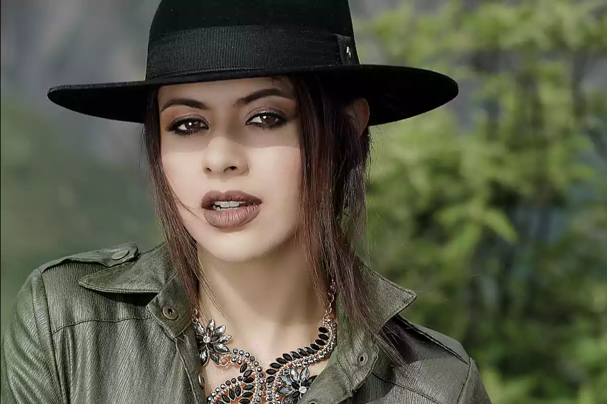 Mujer con sombrero negro, chaqueta verde y collar llamativo posando al aire libre