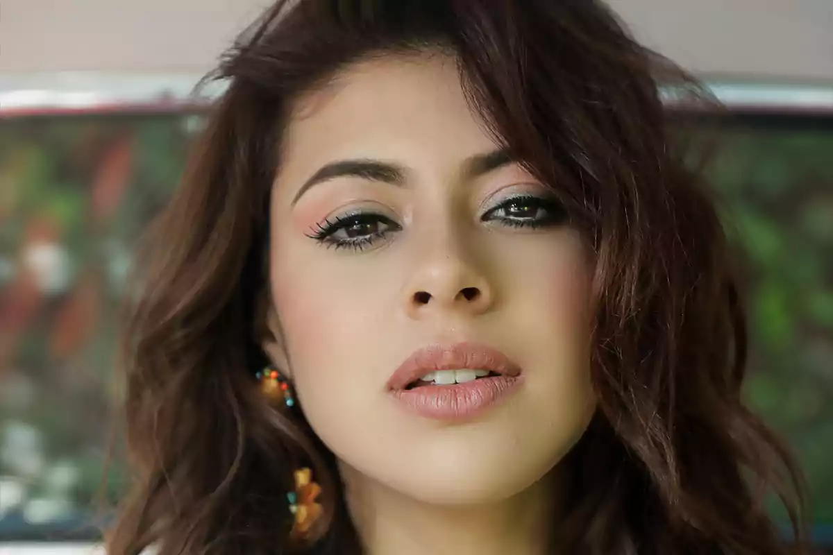 Retrato de una mujer joven con maquillaje natural y labios nude, cabello castaño suelto y pendientes coloridos, fondo desenfocado con tonos verdes y rojizos. Retrato de una mujer joven con maquillaje natural y labios nude, cabello castaño suelto y pendientes coloridos, fondo desenfocado con tonos verdes y rojizos.