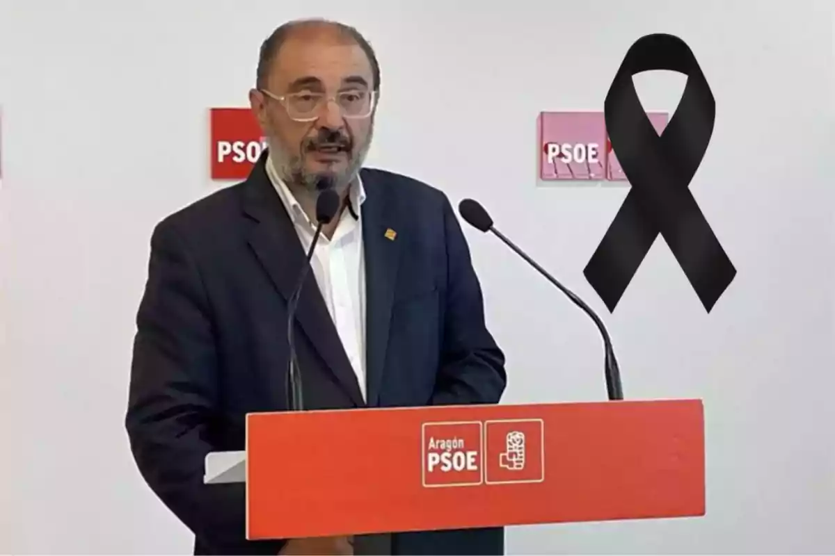 Hombre con gafas y barba hablando en un atril del PSOE Aragón con un lazo negro de luto al fondo