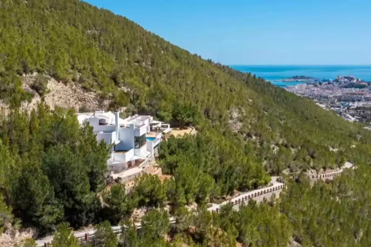 Casa blanca moderna rodeada de árboles en una ladera con vista al mar y a una ciudad costera a lo lejos Casa blanca moderna rodeada de árboles en una ladera con vista al mar y a una ciudad costera a lo lejos