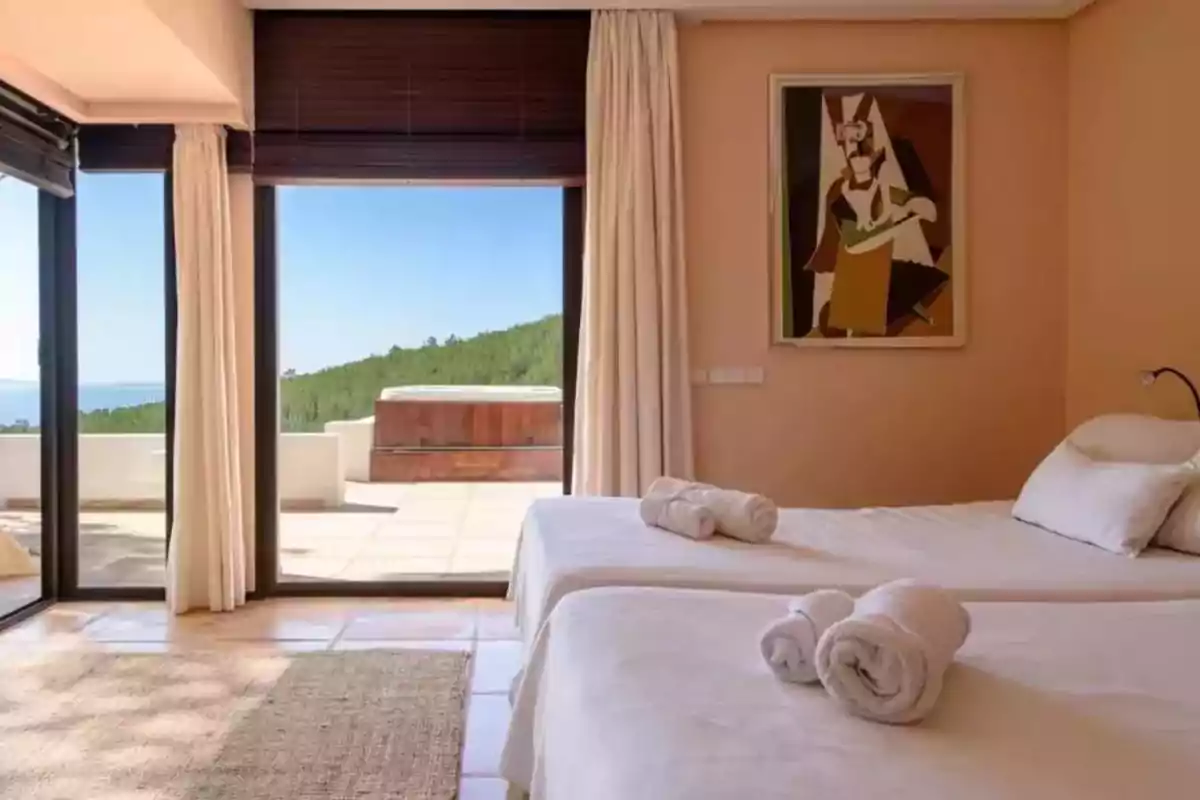 Habitación luminosa con dos camas individuales, toallas enrolladas sobre las camas, grandes ventanales con vista a una terraza y jacuzzi exterior, paredes color durazno y un cuadro moderno en la pared Habitación luminosa con dos camas individuales, toallas enrolladas sobre las camas, grandes ventanales con vista a una terraza y jacuzzi exterior, paredes color durazno y un cuadro moderno en la pared