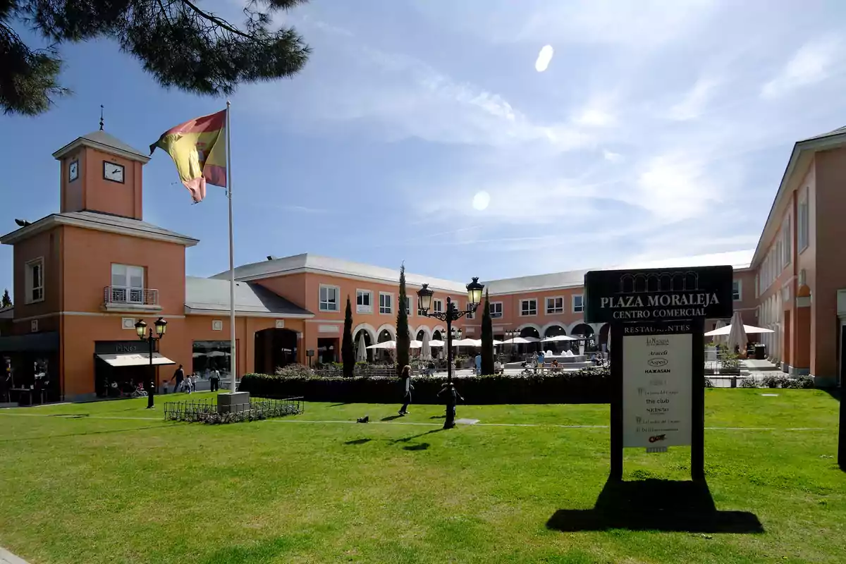 Centro comercial al aire libre con edificios de ladrillo, una gran bandera de España ondeando y un cartel que indica Plaza Moraleja, rodeado de césped y personas paseando en un día soleado. Centro comercial al aire libre con edificios de ladrillo, una gran bandera de España ondeando y un cartel que indica Plaza Moraleja, rodeado de césped y personas paseando en un día soleado.