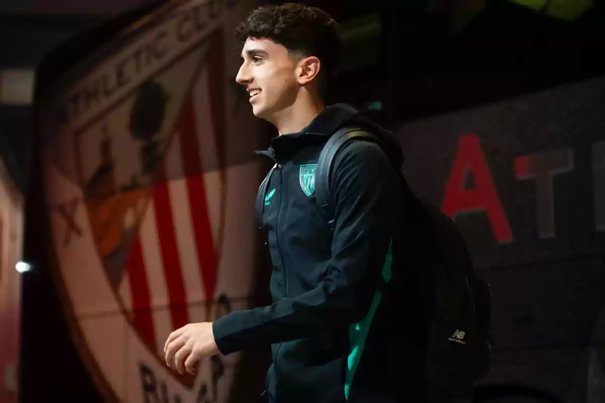 Joven con chaqueta deportiva y mochila sonriendo mientras camina frente a un autobús del Athletic Club Joven con chaqueta deportiva y mochila sonriendo mientras camina frente a un autobús del Athletic Club