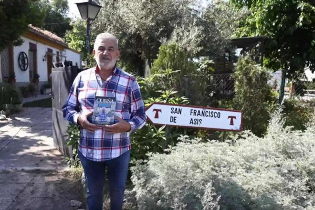 Hombre de cabello canoso y barba con camisa de cuadros sosteniendo un libro frente a un jardín y un letrero que indica San Francisco de Asís Hombre de cabello canoso y barba con camisa de cuadros sosteniendo un libro frente a un jardín y un letrero que indica San Francisco de Asís