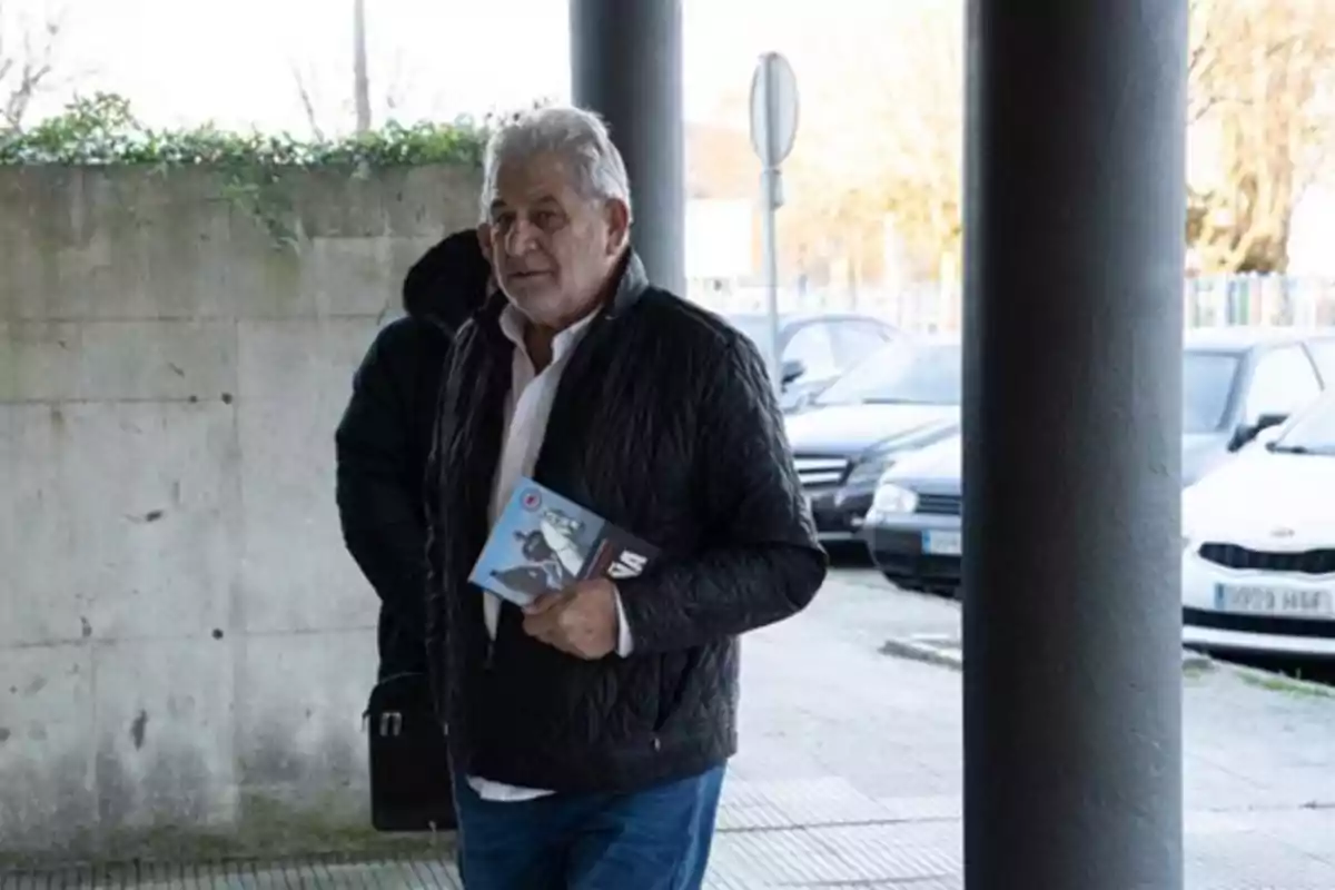 Hombre de cabello canoso con chaqueta negra y camisa blanca caminando al aire libre mientras sostiene un libro en la mano Hombre de cabello canoso con chaqueta negra y camisa blanca caminando al aire libre mientras sostiene un libro en la mano