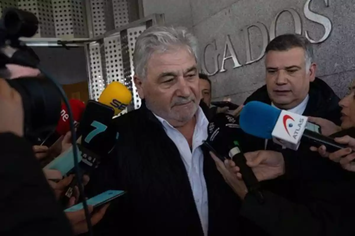 Un hombre de cabello canoso es rodeado por periodistas y micrófonos a la salida de un edificio con la palabra "JUZGADOS" en la pared.