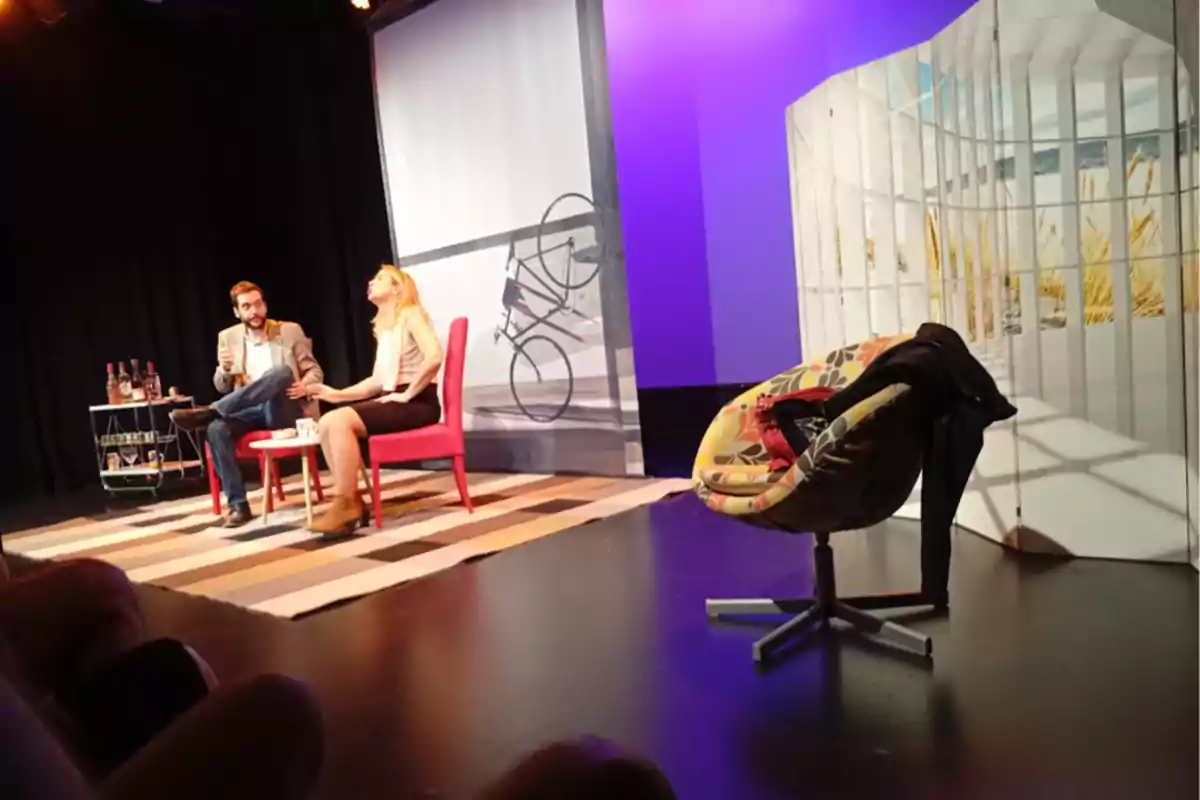 Dos personas sentadas en sillas rojas conversan en un escenario de teatro con fondo de bicicleta y una silla colorida a la derecha Dos personas sentadas en sillas rojas conversan en un escenario de teatro con fondo de bicicleta y una silla colorida a la derecha