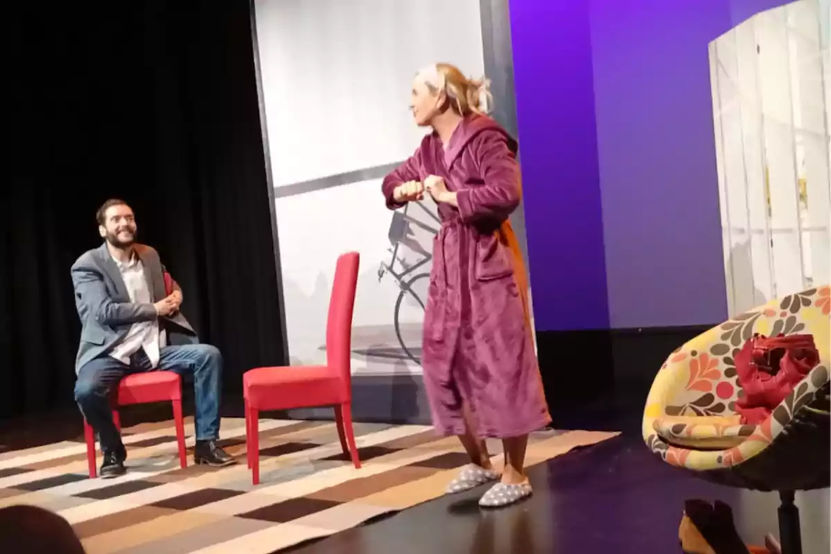 Dos personas actúan en una obra de teatro, una mujer con bata y pantuflas está de pie gesticulando mientras un hombre con saco y jeans la observa sentado en una silla roja, el escenario tiene una alfombra a cuadros y una silla con estampado colorido. Dos personas actúan en una obra de teatro, una mujer con bata y pantuflas está de pie gesticulando mientras un hombre con saco y jeans la observa sentado en una silla roja, el escenario tiene una alfombra a cuadros y una silla con estampado colorido.