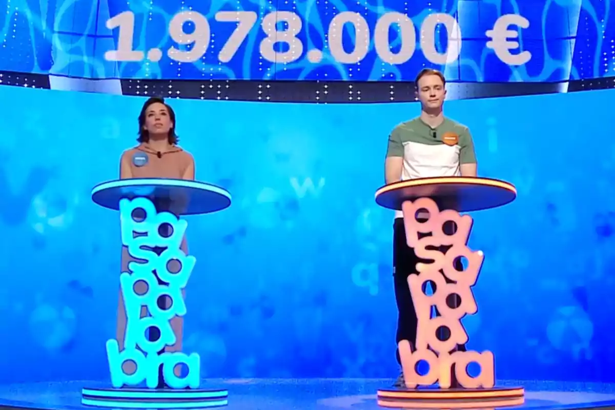 Rosa y Manu de pie en un plató de televisión con decoración azul y naranja, cada uno detrás de un atril con la palabra Rosa y Manu de pie en un plató de televisión con decoración azul y naranja, cada uno detrás de un atril con la palabra