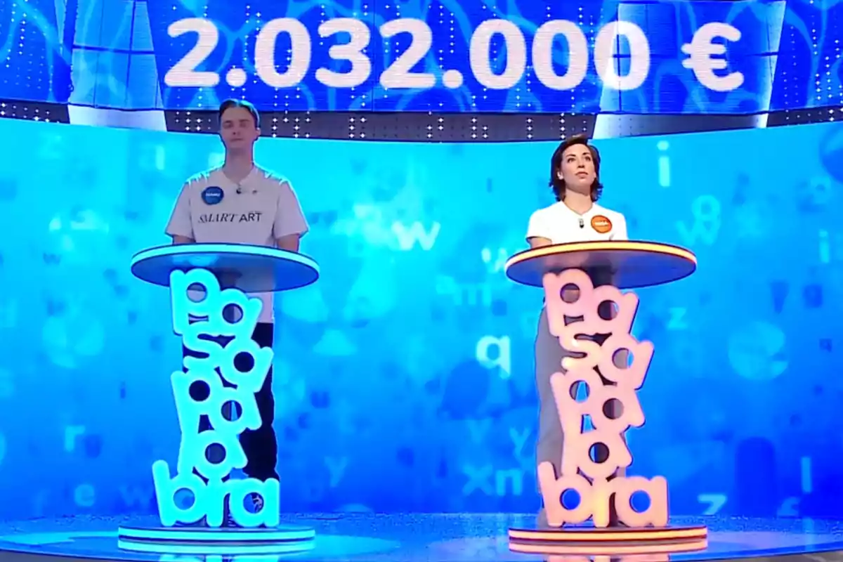 Manu y Rosa de pie detrás de atriles iluminados en un plató de televisión con un fondo azul y una cifra grande de dinero en euros en la parte superior. Captura de 'Pasapalabra' Manu y Rosa de pie detrás de atriles iluminados en un plató de televisión con un fondo azul y una cifra grande de dinero en euros en la parte superior. Captura de 'Pasapalabra'