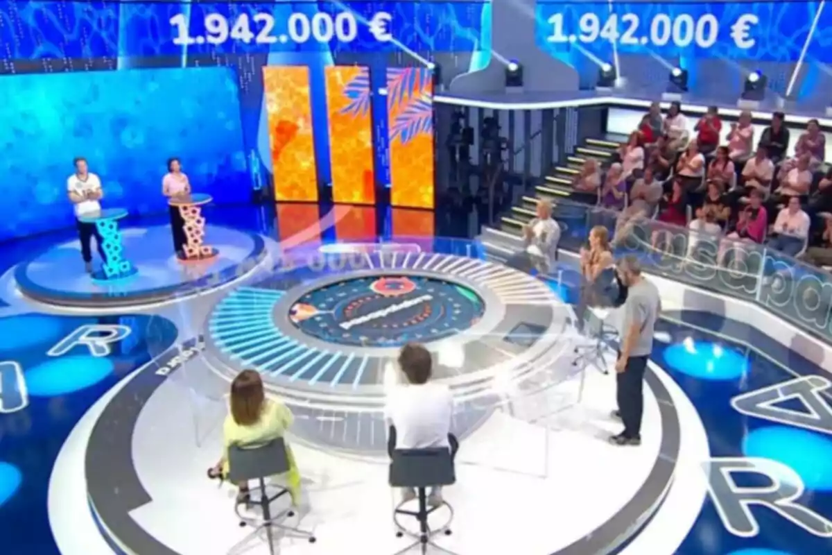 Un plató de concurso televisivo con dos concursantes de pie frente a atriles, un panel circular en el centro, público sentado al fondo y una pantalla que muestra una cantidad de dinero en euros. Captura de 'Pasapalabra' Un plató de concurso televisivo con dos concursantes de pie frente a atriles, un panel circular en el centro, público sentado al fondo y una pantalla que muestra una cantidad de dinero en euros. Captura de 'Pasapalabra'