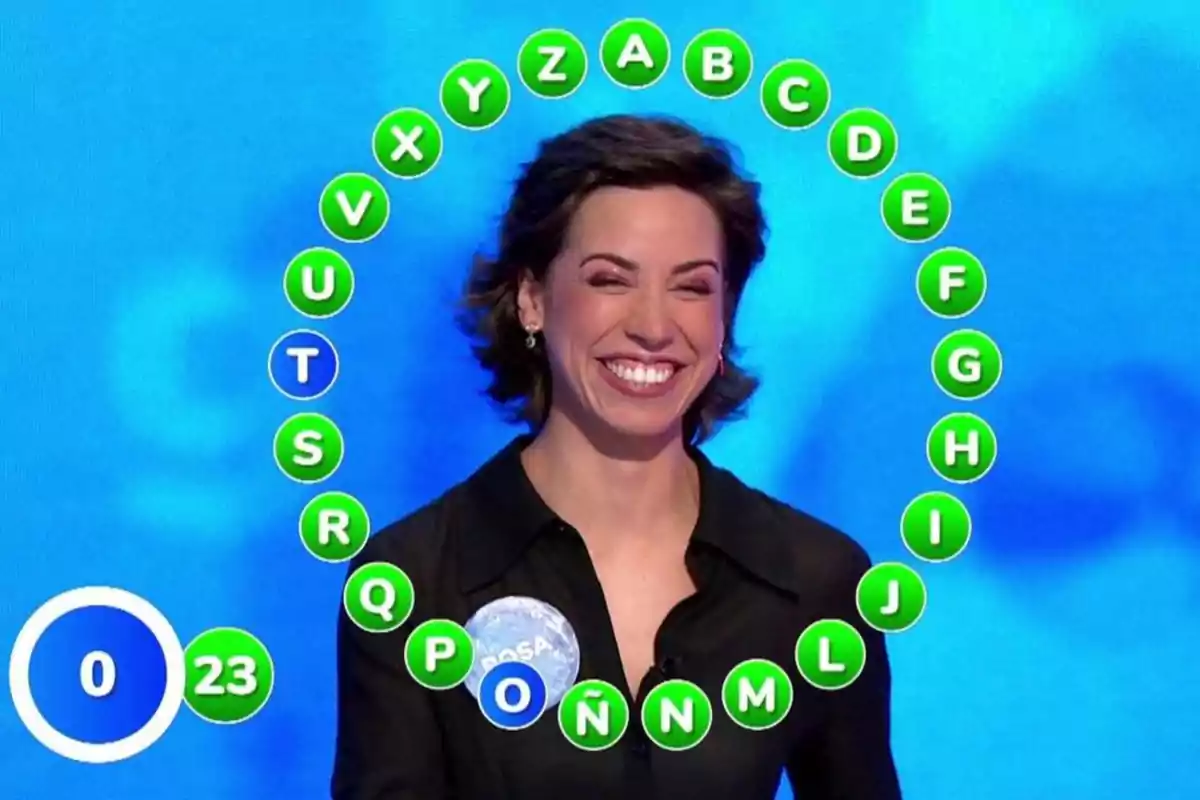 Rosa Rodríguez sonriente participando en un concurso de televisión con letras del abecedario en círculos verdes formando un semicírculo a su alrededor y un marcador con el número 23. Captura de 'Pasapalabra' Rosa Rodríguez sonriente participando en un concurso de televisión con letras del abecedario en círculos verdes formando un semicírculo a su alrededor y un marcador con el número 23. Captura de 'Pasapalabra'
