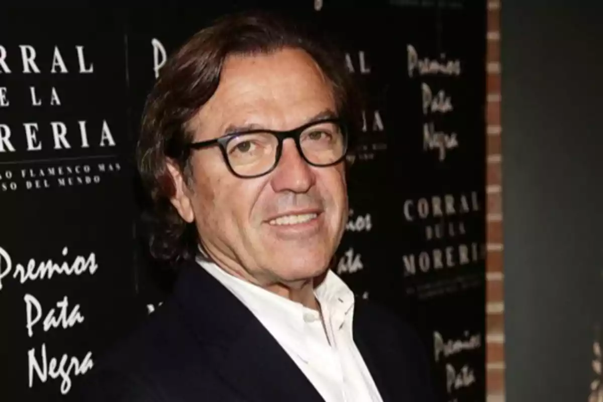 Hombre de cabello castaño y gafas con saco oscuro y camisa blanca posando frente a un fondo con letras blancas