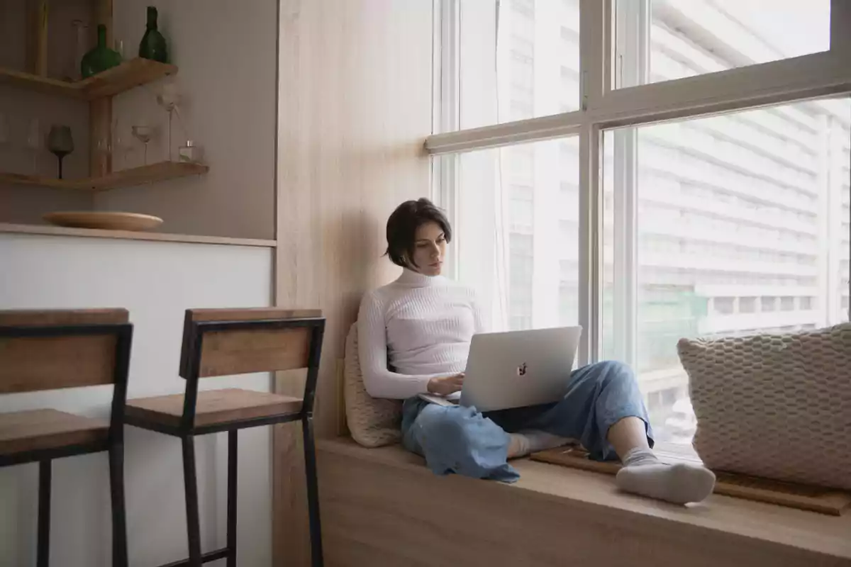 Mujer sentada junto a una ventana trabajando en una laptop en un ambiente hogareño y moderno