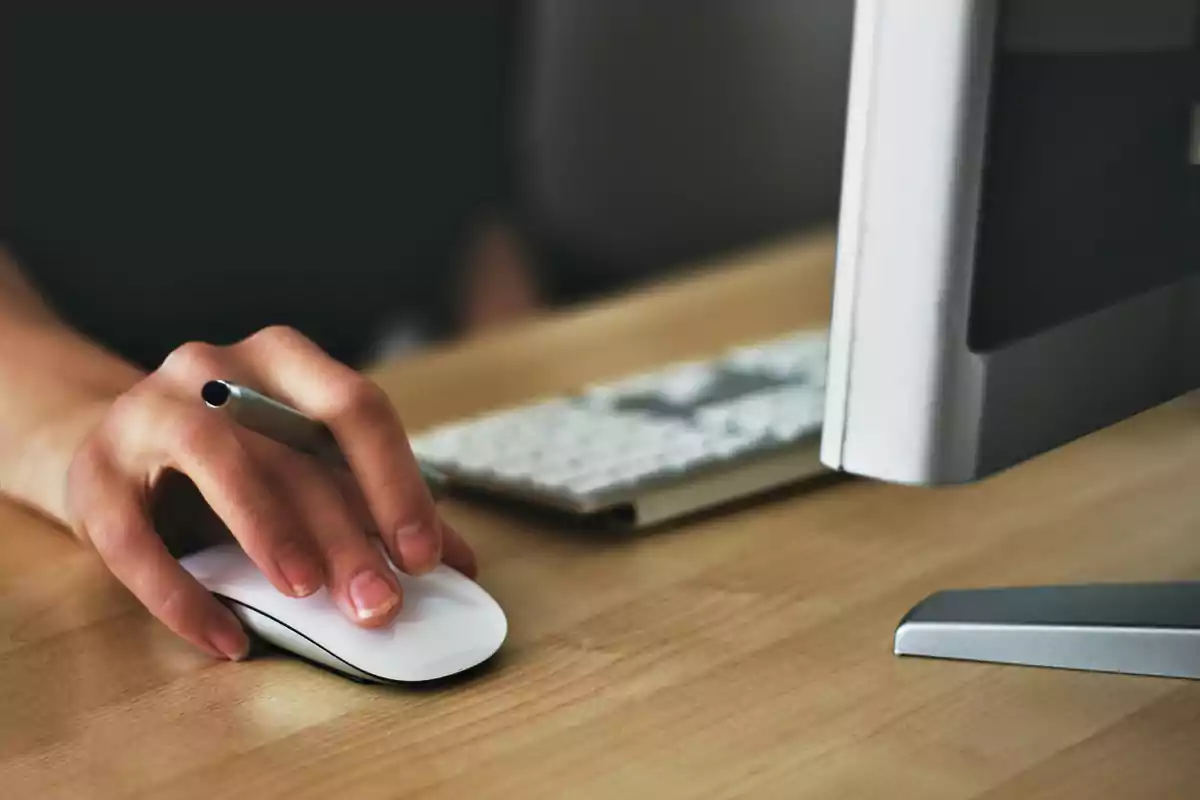 Persona usando un mouse de computadora junto a un teclado y monitor sobre un escritorio de madera