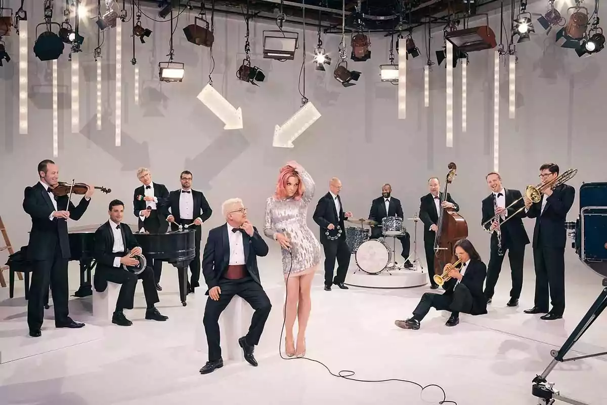 Un grupo de músicos elegantemente vestidos toca diferentes instrumentos junto a una cantante con vestido plateado en un estudio iluminado
