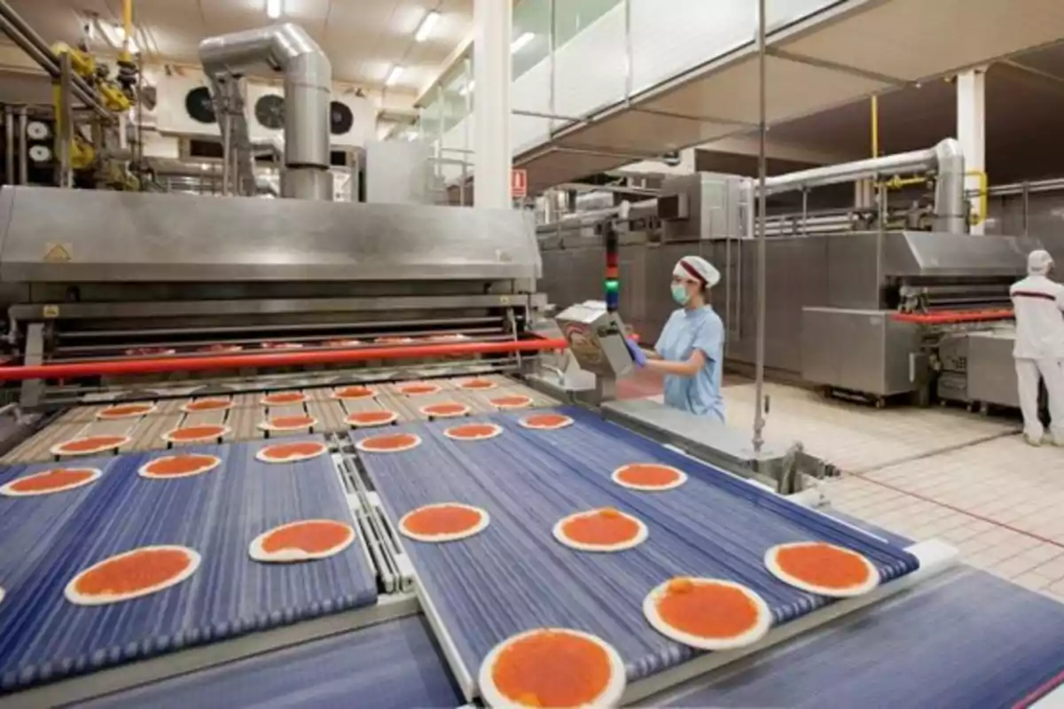Línea de producción industrial de pizzas con trabajadores usando uniforme y mascarilla supervisando el proceso Línea de producción industrial de pizzas con trabajadores usando uniforme y mascarilla supervisando el proceso