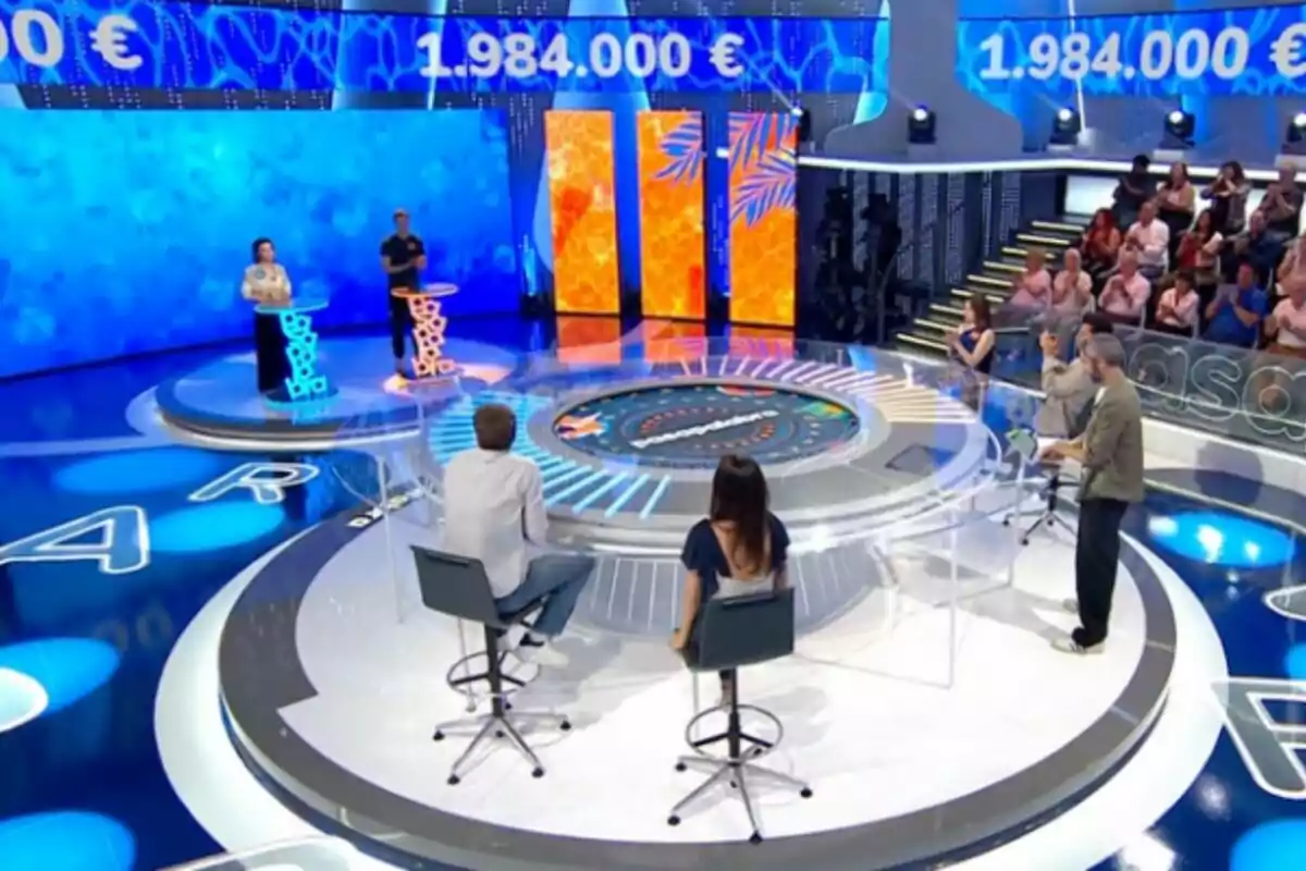 Un plató de concurso televisivo, que es 'Pasapalabra', con dos concursantes de pie en atriles, dos personas sentadas de espaldas y un presentador de pie, con público al fondo y una pantalla que muestra una cantidad de dinero. Un plató de concurso televisivo, que es 'Pasapalabra', con dos concursantes de pie en atriles, dos personas sentadas de espaldas y un presentador de pie, con público al fondo y una pantalla que muestra una cantidad de dinero.