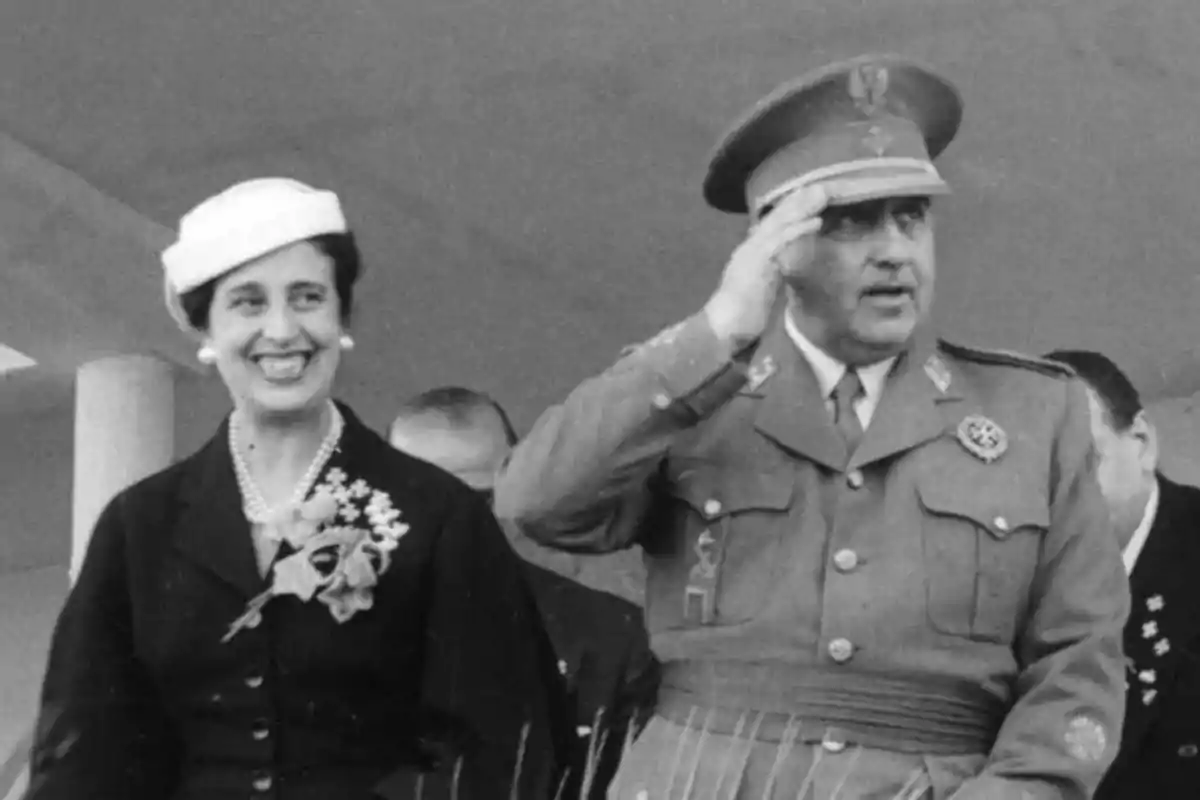 Una mujer sonriente con sombrero blanco y un hombre con uniforme militar saludando, ambos en un evento público antiguo. Una mujer sonriente con sombrero blanco y un hombre con uniforme militar saludando, ambos en un evento público antiguo.