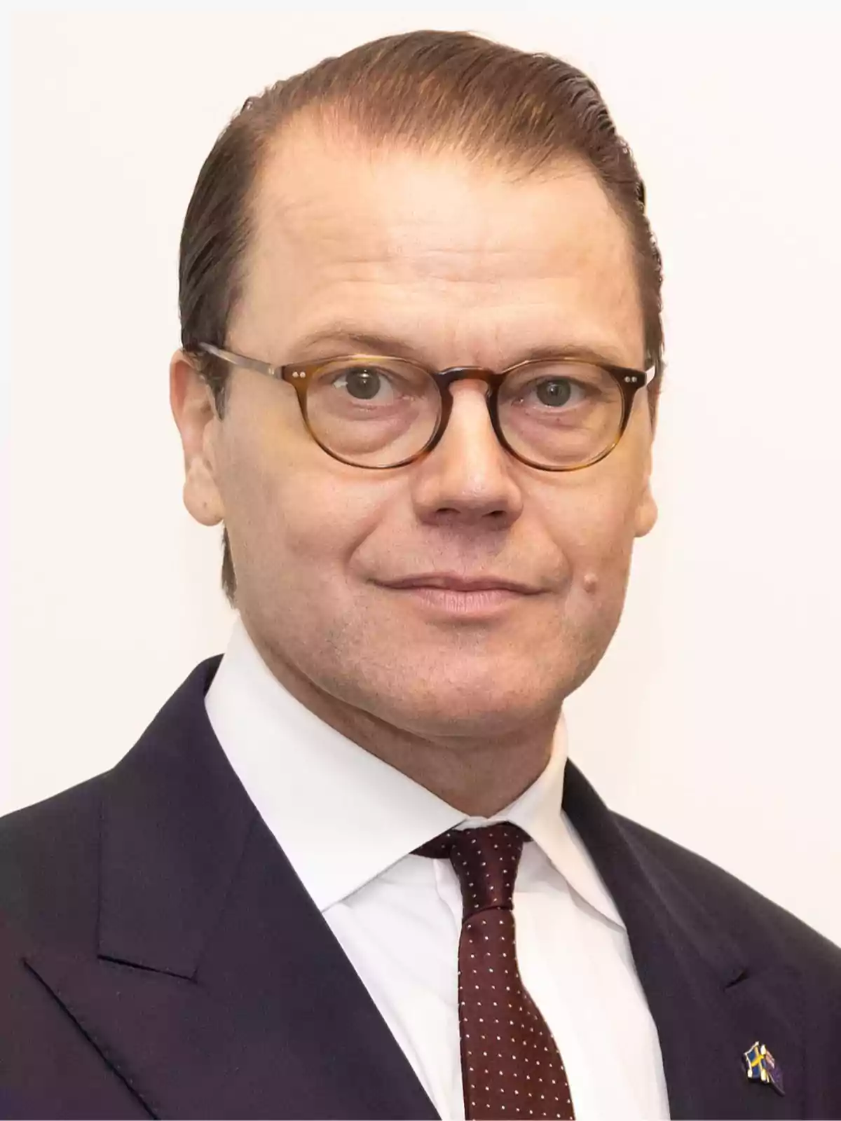 Hombre de cabello castaño peinado hacia atrás, con gafas redondas, traje oscuro, camisa blanca y corbata marrón con puntos blancos, posando frente a un fondo claro. Hombre de cabello castaño peinado hacia atrás, con gafas redondas, traje oscuro, camisa blanca y corbata marrón con puntos blancos, posando frente a un fondo claro.