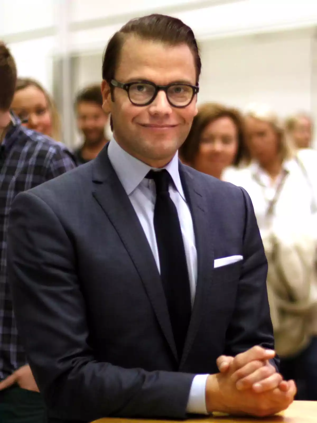 Hombre con gafas y traje oscuro sonriendo mientras está de pie con las manos entrelazadas en un entorno interior con personas al fondo desenfocadas Hombre con gafas y traje oscuro sonriendo mientras está de pie con las manos entrelazadas en un entorno interior con personas al fondo desenfocadas