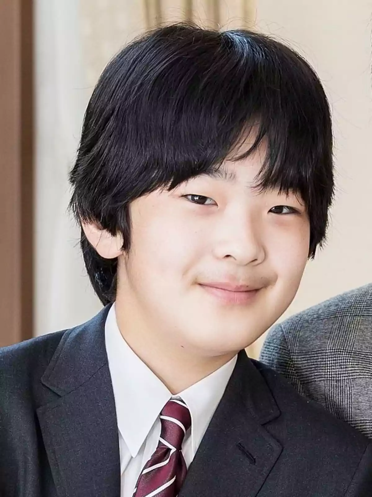 Joven de cabello negro y lacio vistiendo traje oscuro, camisa blanca y corbata a rayas, sonriendo levemente frente a un fondo interior claro Joven de cabello negro y lacio vistiendo traje oscuro, camisa blanca y corbata a rayas, sonriendo levemente frente a un fondo interior claro