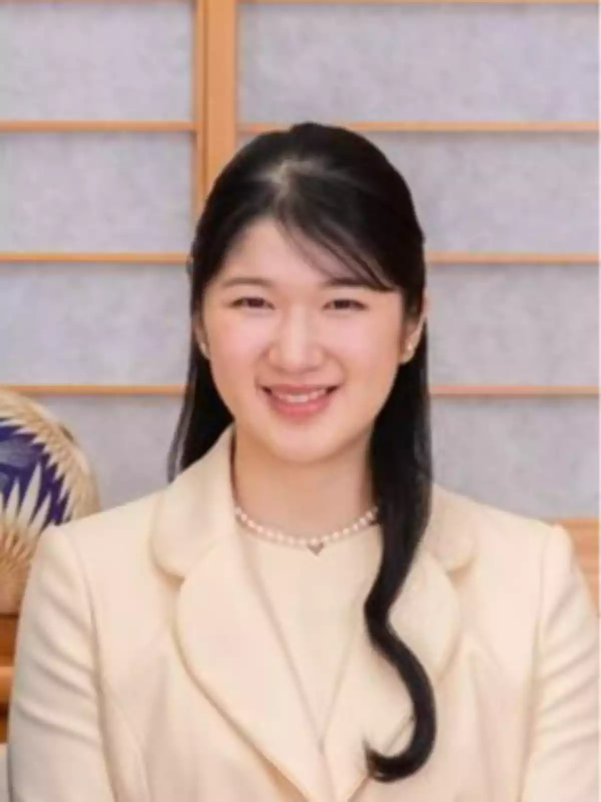 Mujer joven de cabello largo y oscuro vestida con un traje claro y collar de perlas, sonriendo frente a un fondo tradicional japonés. Mujer joven de cabello largo y oscuro vestida con un traje claro y collar de perlas, sonriendo frente a un fondo tradicional japonés.