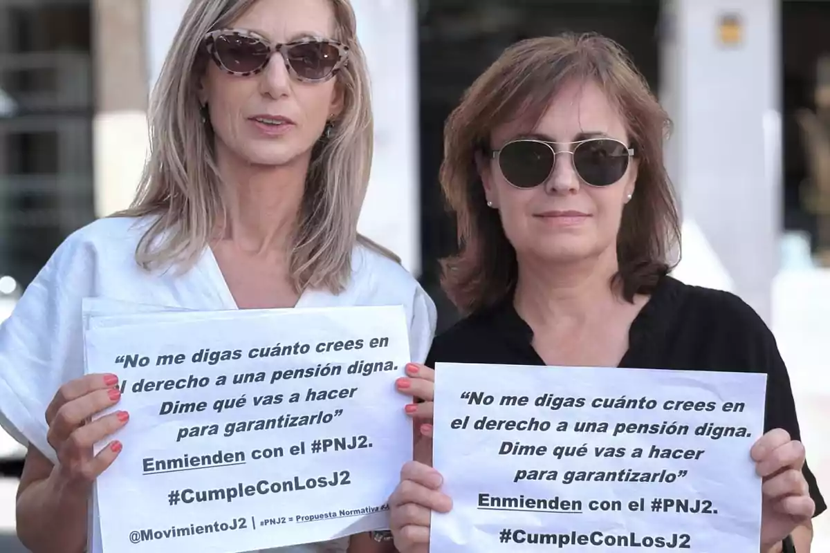 Dos mujeres con gafas de sol sostienen carteles impresos con mensajes en defensa de una pensión digna y el hashtag CumpleConLosJ2 Dos mujeres con gafas de sol sostienen carteles impresos con mensajes en defensa de una pensión digna y el hashtag CumpleConLosJ2